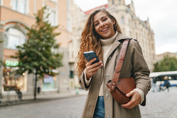 Smiling curly woman wearing warm coat walking down the street and using her phone