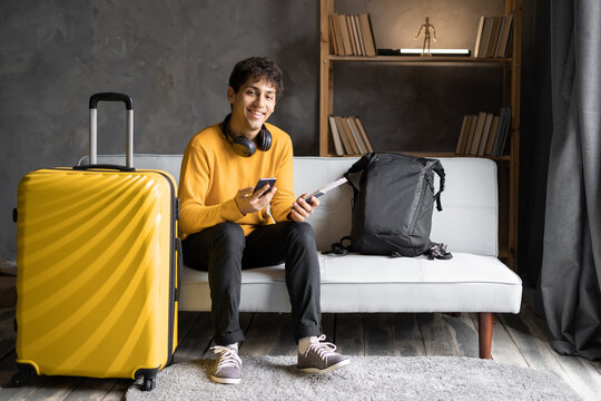 Vacation. Happy traveler young man ordering taxi to airport by smartphone app sitting with suitcase at home, looking at camera.