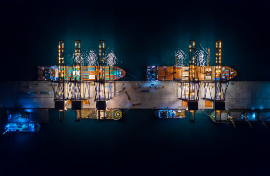 Aerial top view of international port with crane loading containers in import export business logistics. Container loading and unloading cargo freight ship in deep sea at night. - Powered by Adobe