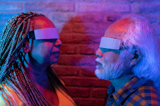 Portrait of senior multiracial couple looking at each other with virtual reality goggles in a neon light room. Focus on mature asian man