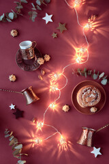 Eid Mubarak winter muslim festival. Flat lay with copper cezve coffee pots, traditional bronze metal cup holder, marzipan sweets, wooden stars and eucalyptus twigs. Dark red flat lay, light garland.