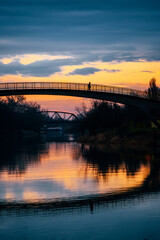 Fototapeta na wymiar Silhouette of a man walking across small bridge over a peaceful river at sunset. Serene scenery in Zrenjanin city in Serbia on the river Begej