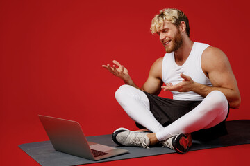Fototapeta na wymiar Full body young strong sporty sportsman man wear white clothes spend time in home gym use laptop pc computer speak talk sit on yoga mat isolated on plain red background Workout sport fit body concept.