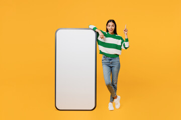 Full body young fun latin woman wear casual cozy green knitted sweater big huge blank screen mobile cell phone smartphone with area point finger up isolated on plain yellow background studio portrait.