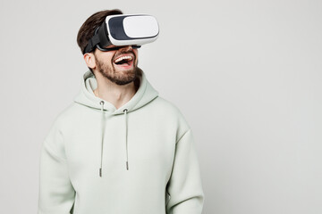 Young smiling amazed happy fun cheerful gambling caucasian man wearing mint hoody watching in vr headset pc gadget isolated on plain solid white background studio portrait. People lifestyle concept.