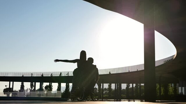 A man in a chair for people with physical disabilities and a woman dancing at sunset.