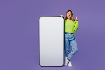 Full body young woman 30s she wear casual green knitted sweater stand near big huge blank screen mobile cell phone with area point index finger up isolated on plain pastel purple background studio.