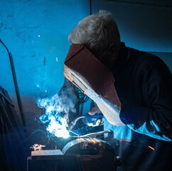 worker, pensioner welding in his workshop. He wears a protective welding mask but does not use protective gloves. Dangerous! Safety and protection at work. The protective mask is old and dusty