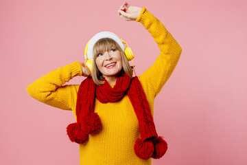 Merry elderly woman 50s year old wear yellow knitted sweater headphones red scarf Santa hat posing listen music raise up hands isolated on plain pink background. Happy New Year Christmas 2023 concept
