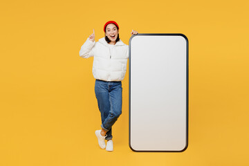 Full body young woman of Asian ethnicity wear white padded windbreaker jacket red hat big huge blank screen mobile cell phone smartphone with area point finger up isolated on plain yellow background.