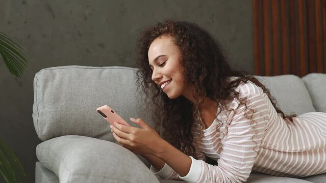 Young happy calm woman wear casual clothes lying on grey sofa couch hold in hand use mobile cell phone chatting put down turn around stay at home hotel flat relax spend free spare time in living room