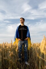 Naklejka na ściany i meble a young guy stands in a wheat field against the background of a blue sky with textured clouds with the Ukrainian flag on his shoulders with his hands in his pockets. Photo at sunset