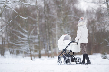Fototapeta na wymiar Young adult mother pushing white baby stroller and walking on snow covered sidewalk at park in cold winter day. Spending time with newborn and breathing fresh air. Enjoying peaceful stroll. Back view.