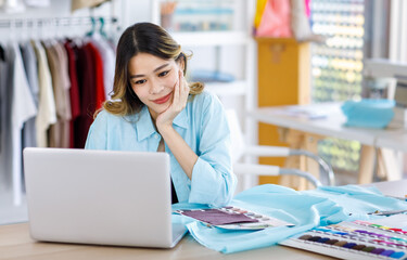 Millennial Asian young beautiful professional female designer dressmaker sitting smiling at working table with clothing color pattern sample and fabric using laptop computer in tailor studio workshop