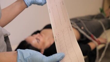 Doctor cardiologist looking on cardiogram of patient. Female hands of professional doctor hold cardiogram for further examination of a sick woman in a modern clinic. Woman lies on blurred background. - Powered by Adobe