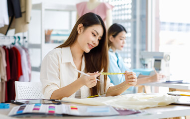 Millennial Asian young beautiful professional female designer dressmaker businesswoman measure fabric by measuring tape on working desk while colleague using sewing machine in tailor workshop studio.