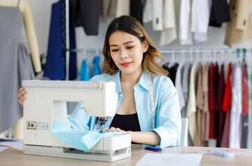 Obraz na płótnie Canvas Millennial Asian young professional female designer dressmaker businesswoman with measuring tape around neck sitting using sewing machine sew fabric textile at working desk in tailor workshop studio