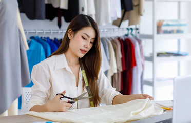 Millennial Asian young professional female designer dressmaker businesswoman with measuring tape using scissors cutting fabric textile on working desk with laptop and tablet in tailor workshop studio