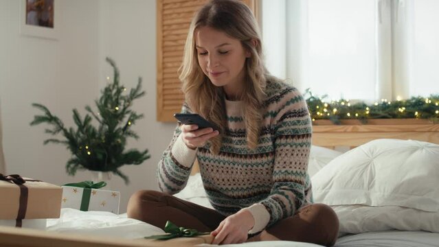 Caucasian woman sitting on bed and taking picture with smart phone of wrapped Christmas presents. Shot with RED helium camera in 8K.  