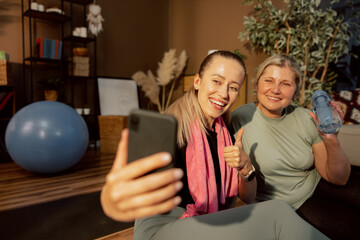 Delighted young mother and senior woman sitting together on yoga may relaxing after difficult yoga lesson talking with offspring by video link. Beautiful girl sowinh thumb up at camera.