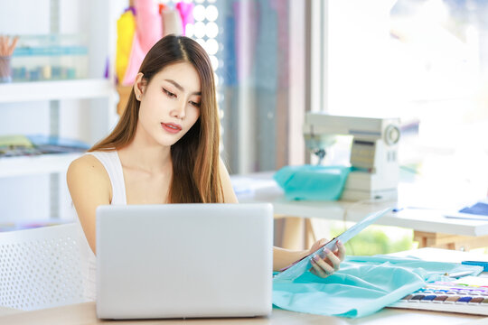 Millennial Asian young professional female dressmaker designer seamstress sitting working alone with laptop computer holding color shading pattern paper collection in tailor workshop studio office