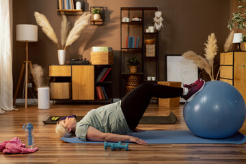 Elderly woman laying in floor mat yoga exercising training with fitball at cozy home indoors using...