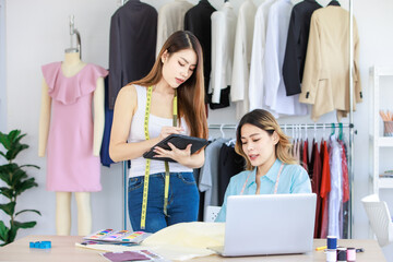 Millennial Asian young professional female dressmaker designer seamstress mentor with measuring tape helping teaching advising colleague choosing color pattern chart in tailor studio workshop office