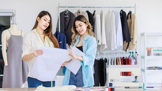 Millennial Asian young professional female dressmaker designer seamstress with measuring tape and colleague partner help checking cutting dress in front clothesline in tailor studio workshop office - Powered by Adobe