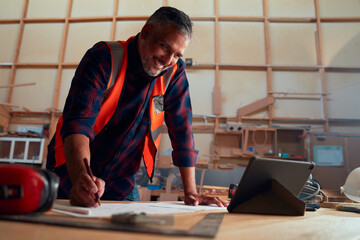 Mid adult man smiling while writing on paper next to digital tablet in woodworking factory