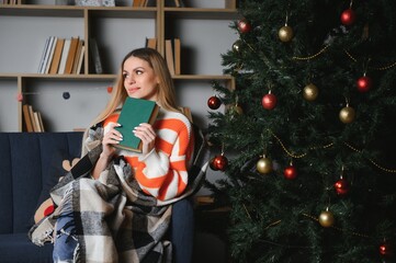 Attractive young lady with dark hair reading interesting book while sitting on grey couch. Blur background of beautiful christmas tree. Cozy atmosphere.