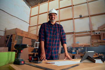 Mid adult man in safety helmet smiling near digital tablet and power tools in woodworking factory