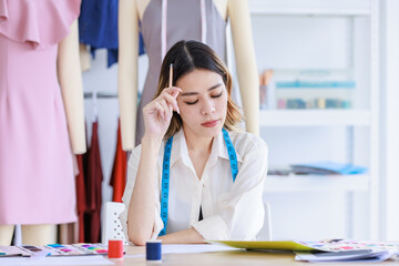 Millennial Asian young thoughtful female professional dressmaker designer seamstress with measuring tape holding pencil sitting working thinking creating ideas at tailor studio workshop office desk