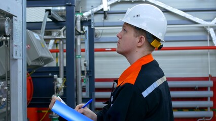 Professional Heavy Industry Engineer or Worker Wearing Safety Uniform and Hard Hat. A young engineer in a white helmet makes entries in a journal about the operation of industrial equipment.