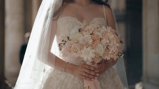 Elegant brunette bride in fashion lace wedding dress and long satin veil standing against blurred groom among the marble columns background, affectionate woman holding charming pastel colours wedding