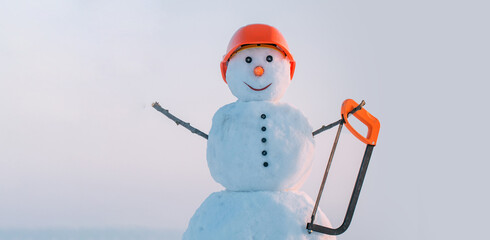 Snowman builder in building helmet hold saw. Snowman in hard hat on the snow outdoor background. Christmas banner with snowman. Snowman ready for building and repair work.