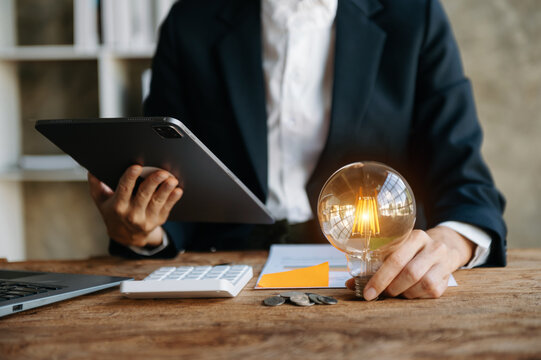 business hand holding lightbulb with using laptop computer and money stack in office. idea saving energy and accounting finance concept  in morning light..