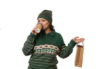 Pretty woman in woolen sweater and hat, drinking hot tea or coffee from a paper cup, holding a...