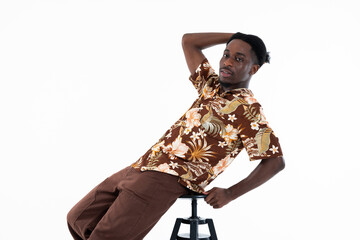 African american man funny posing on chair on white background in studio isolated looking at camera. Wearing colored shirt printed with flowers and brown pants.