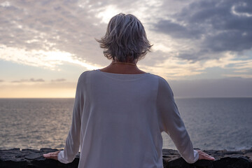 Fototapeta premium Rear view of senior woman standing in front of the sea at sunset looking at the horizon over the water. Peace, solitude, relaxation, meditation
