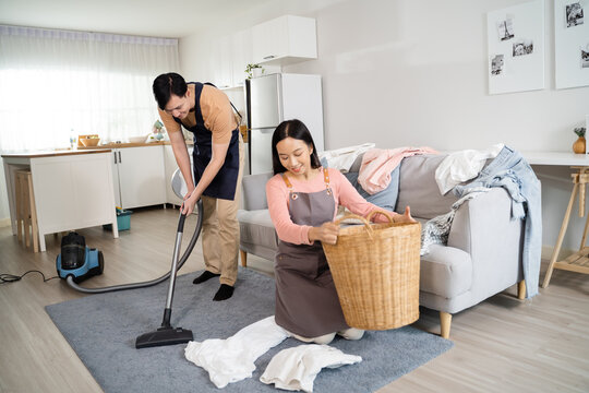 Young Asian Couple Doing Houserkeeping Together At Home. Beautiful Wife Doing Laundry And Husband Doing Vacuum Cleaning On The Floor. Housework And Chores Concept