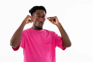 Young man standing on white background in studio looking at camera wearing pink t-shirt and pink glasses holding arms near face posing for fashionable magazine.
