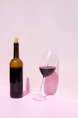 A bottle of red wine and a filled glass next to it in the sun on a pink background. Place for text