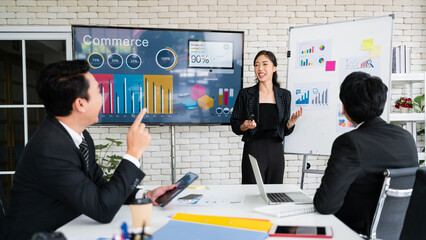 A cheerful and confident Asian businesswoman stands, present bar charts data from a whiteboard to her office colleagues. Asian business women leader role at the meeting.