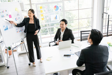 A cheerful and confident Asian businesswoman stands, present bar charts data from a whiteboard to her office colleagues. Asian business women leader role at the meeting.