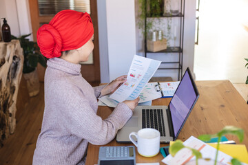 Happy biracial woman wearing hijab, sitting at table in living room, using laptop - Powered by Adobe