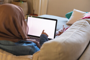 Biracial woman wearing hijab, laying on sofa in living room and using laptop with copy space