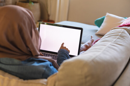 Biracial woman wearing hijab, laying on sofa in living room and using laptop with copy space - Powered by Adobe