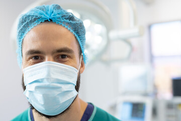 Fototapeta na wymiar Portrait of smiling caucasian male surgeon in surgical mask and cap in operating theatre, copy space