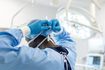 African american male surgeon in surgical gown and cap tying on mask in operating theatre, side view - Powered by Adobe