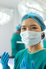 Vertical of smiling asian female surgeon in gown, mask, gloves and cap in theatre, copy space